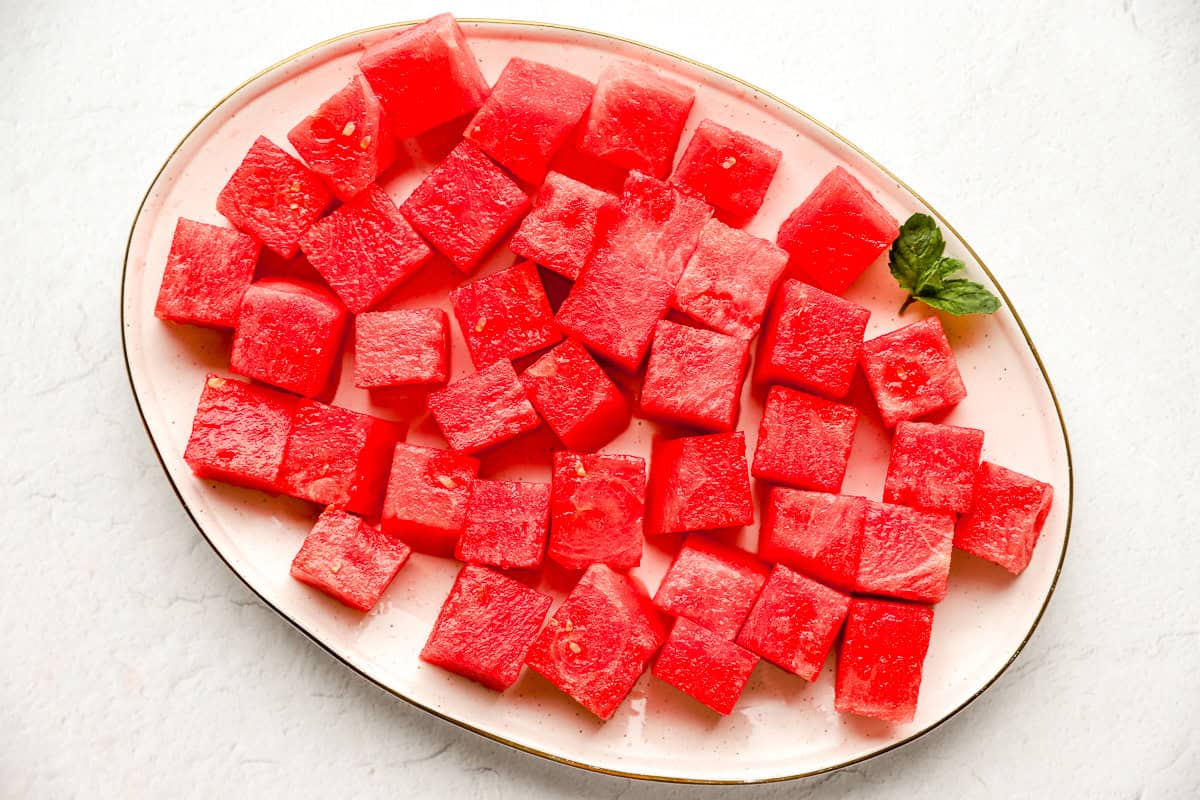 Finished cubes of watermelon neatly arranged on a plate with a sprig of mint for garnish.