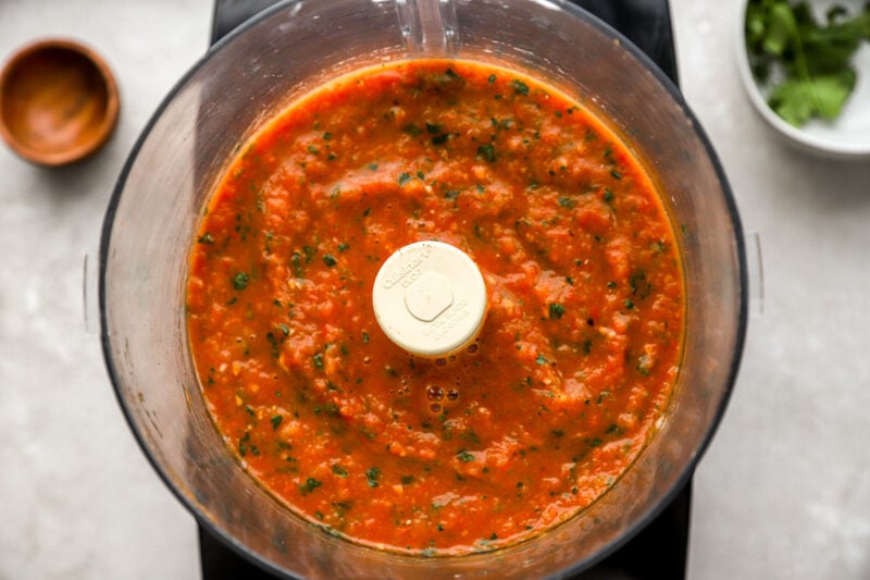 Final blended salsa roja in a food processor, with visible flecks of cilantro and a smooth yet chunky texture.
