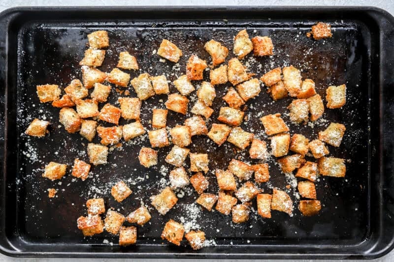 Golden baked croutons on a dark baking sheet, coated with Parmesan cheese.