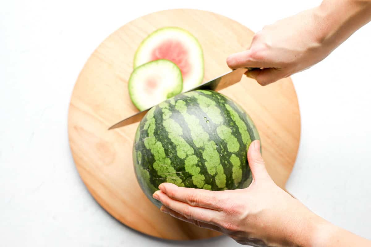 Hands using a knife to slice the top off a whole watermelon on a round wooden cutting board.