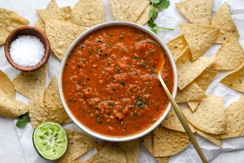 Bowl of freshly made salsa roja with a spoon, surrounded by tortilla chips, lime, cilantro, and a small bowl of salt.