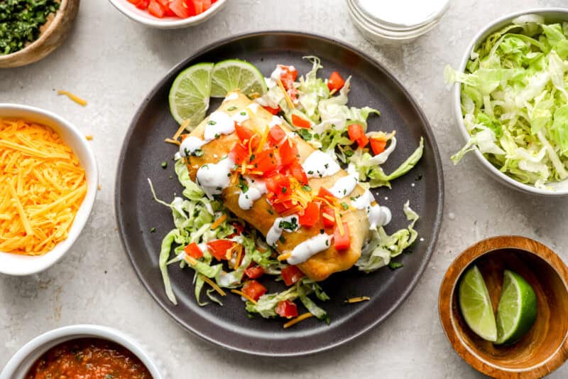 A fully assembled chimichanga served over shredded lettuce and topped with sour cream, diced tomatoes, shredded cheese, and lime wedges, surrounded by bowls of toppings.