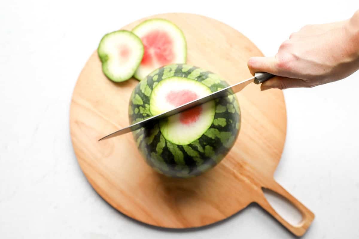 Knife slicing the bottom off a watermelon, revealing the red center, with two rind slices on the side.