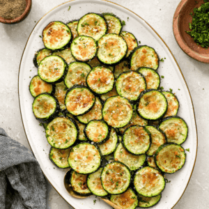 Top-down view of a large plate filled with air fryer zucchini rounds, garnished with Parmesan and parsley, with small bowls of seasoning nearby.