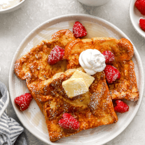 Plate of golden brown brioche French toast topped with raspberries, whipped cream, butter, and powdered sugar.