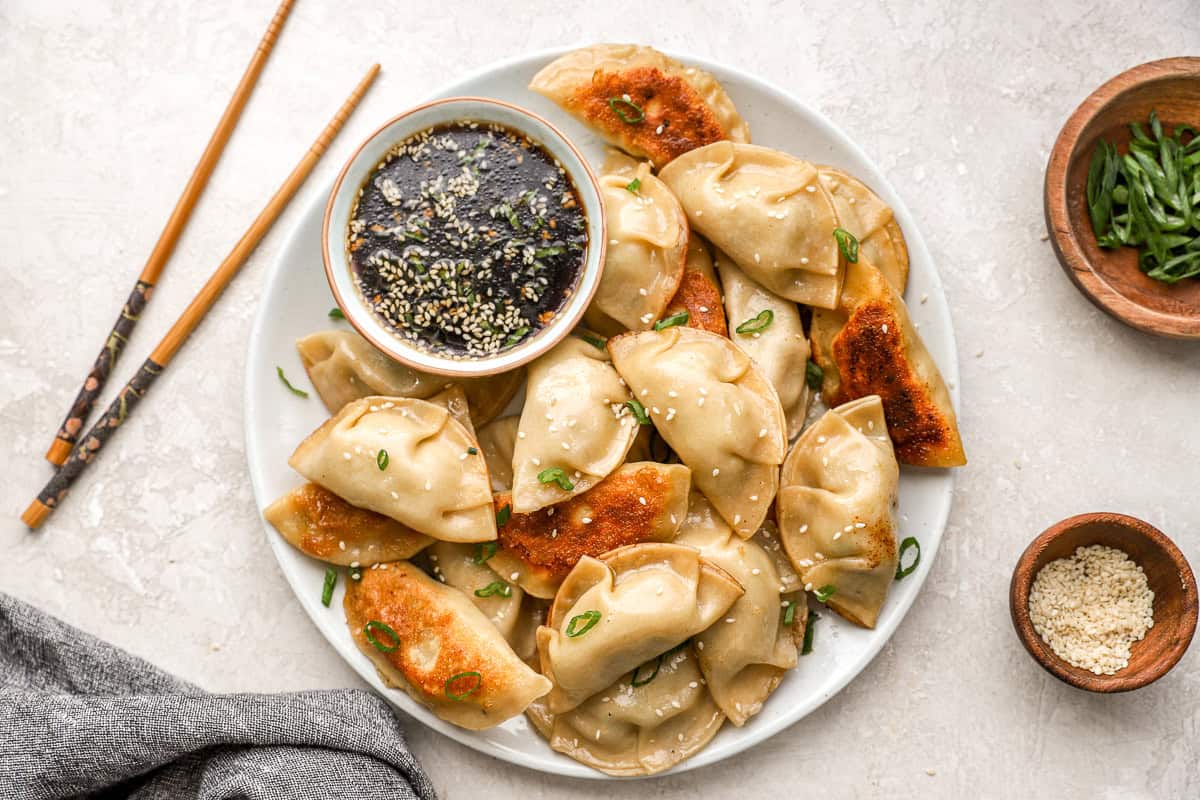A large plate of crispy chicken potstickers garnished with green onions and sesame seeds, served with a small bowl of dipping sauce and chopsticks on the side.