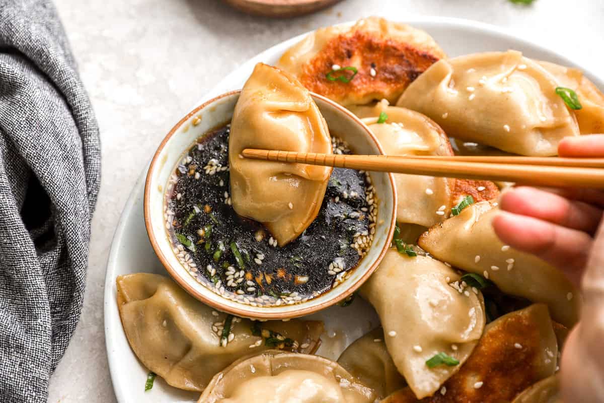 A close-up of a hand holding chopsticks, dipping a glossy chicken potsticker into a bowl of dark soy-based dipping sauce garnished with sesame seeds and chopped green onions. Other golden pan-fried potstickers are arranged around the bowl on a white plate.