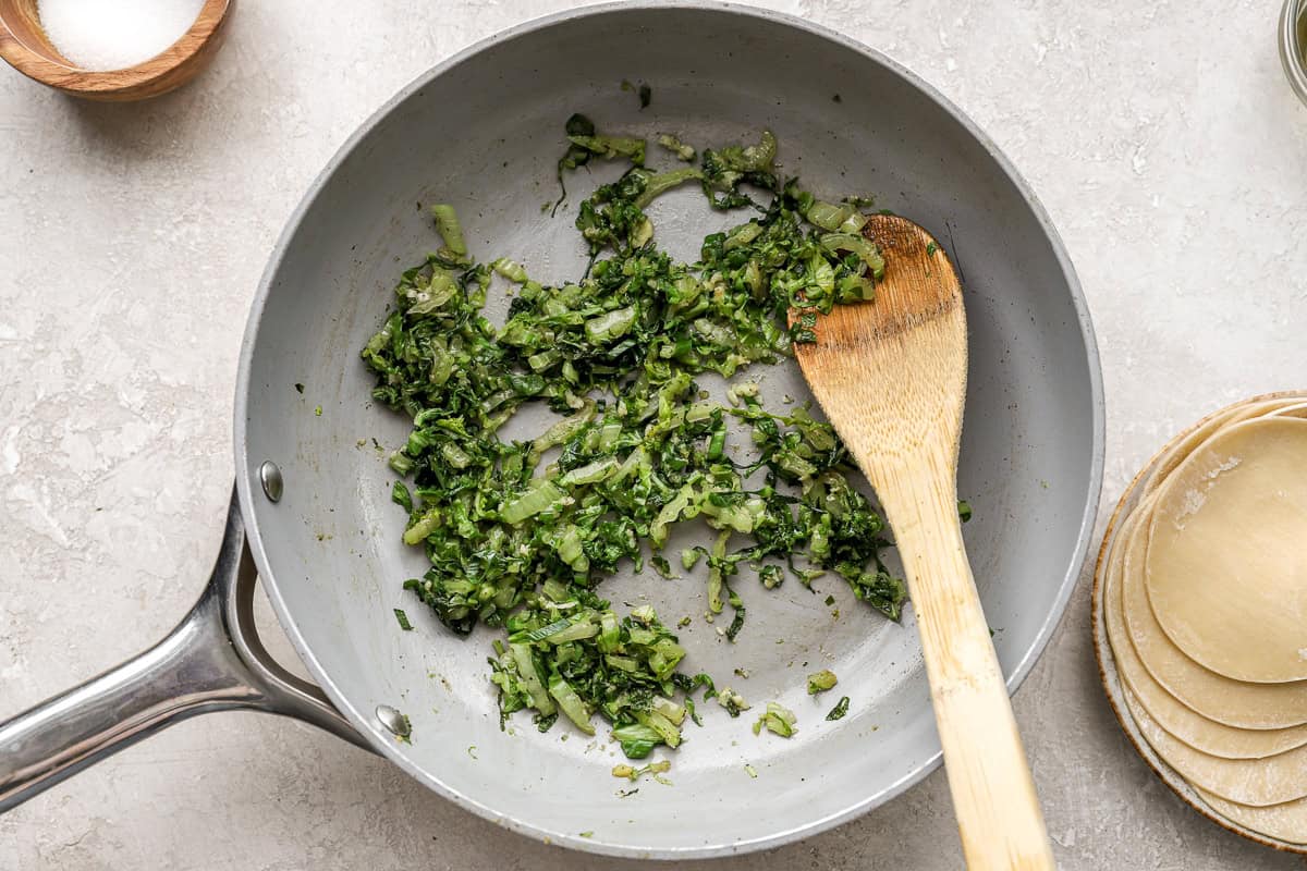 Cabbage, green onions, garlic, and ginger being sautéed in a skillet with a wooden spoon.