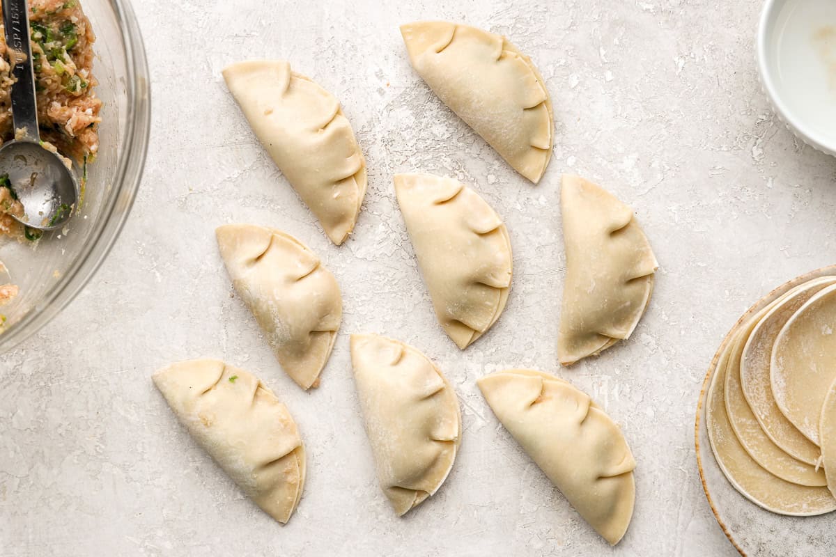 A batch of uncooked chicken potstickers neatly sealed and arranged on a flat surface next to a bowl of water and remaining wrappers.
