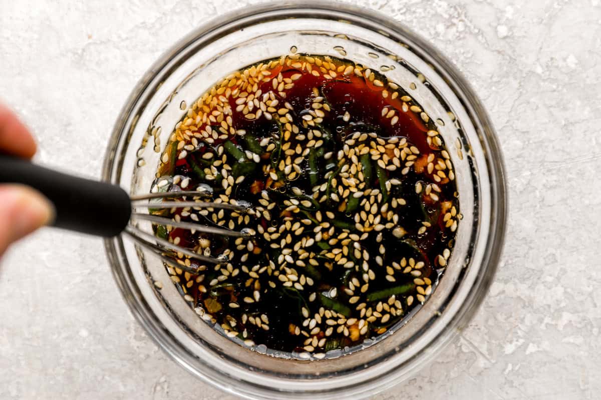 A whisk mixing soy sauce, sesame seeds, chopped green onion, and garlic in a bowl to make the dipping sauce.