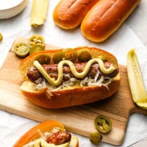 Close-up of a bratwurst in a toasted bun topped with sauerkraut, jalapeños, and mustard, served on a wooden board with pickles and buns in the background.
