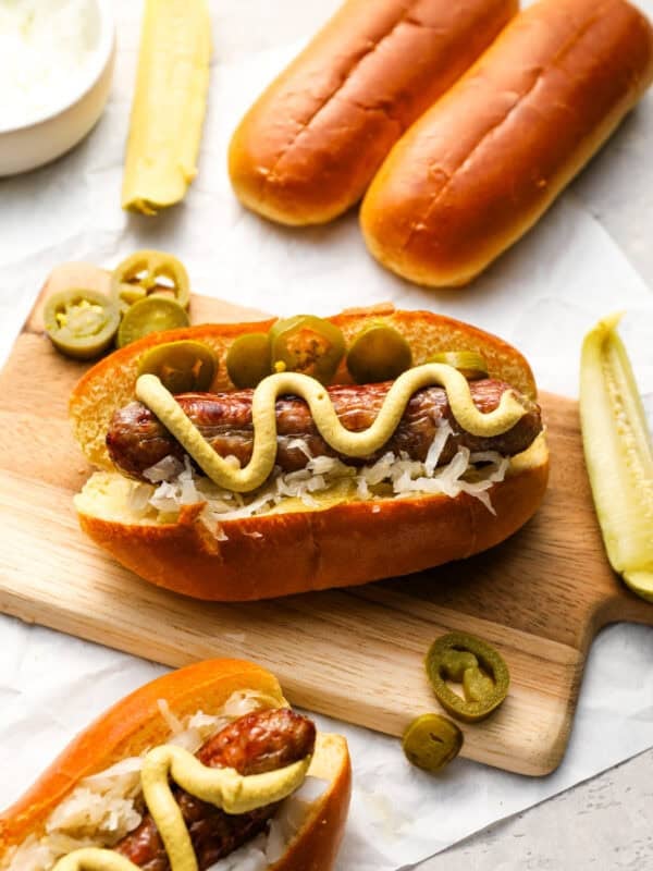 Close-up of a bratwurst in a toasted bun topped with sauerkraut, jalapeños, and mustard, served on a wooden board with pickles and buns in the background.