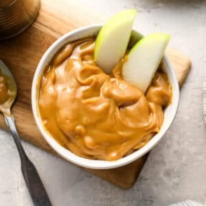 A white bowl filled with thick, glossy caramel and two green apple slices dipped in. The bowl sits on a wooden board next to an opened can of caramel and a spoon.