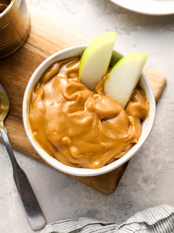 A white bowl filled with thick, glossy caramel and two green apple slices dipped in. The bowl sits on a wooden board next to an opened can of caramel and a spoon.
