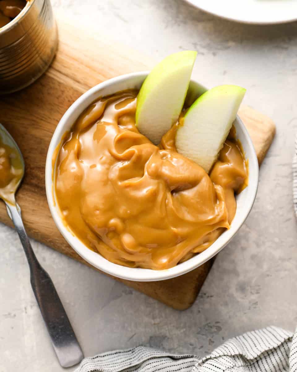 A white bowl filled with thick, glossy caramel and two green apple slices dipped in. The bowl sits on a wooden board next to an opened can of caramel and a spoon.