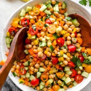 A large white bowl of colorful chickpea salad being mixed with wooden salad servers. The salad features chickpeas, chopped cucumber, halved cherry tomatoes, diced bell peppers, and fresh herbs.