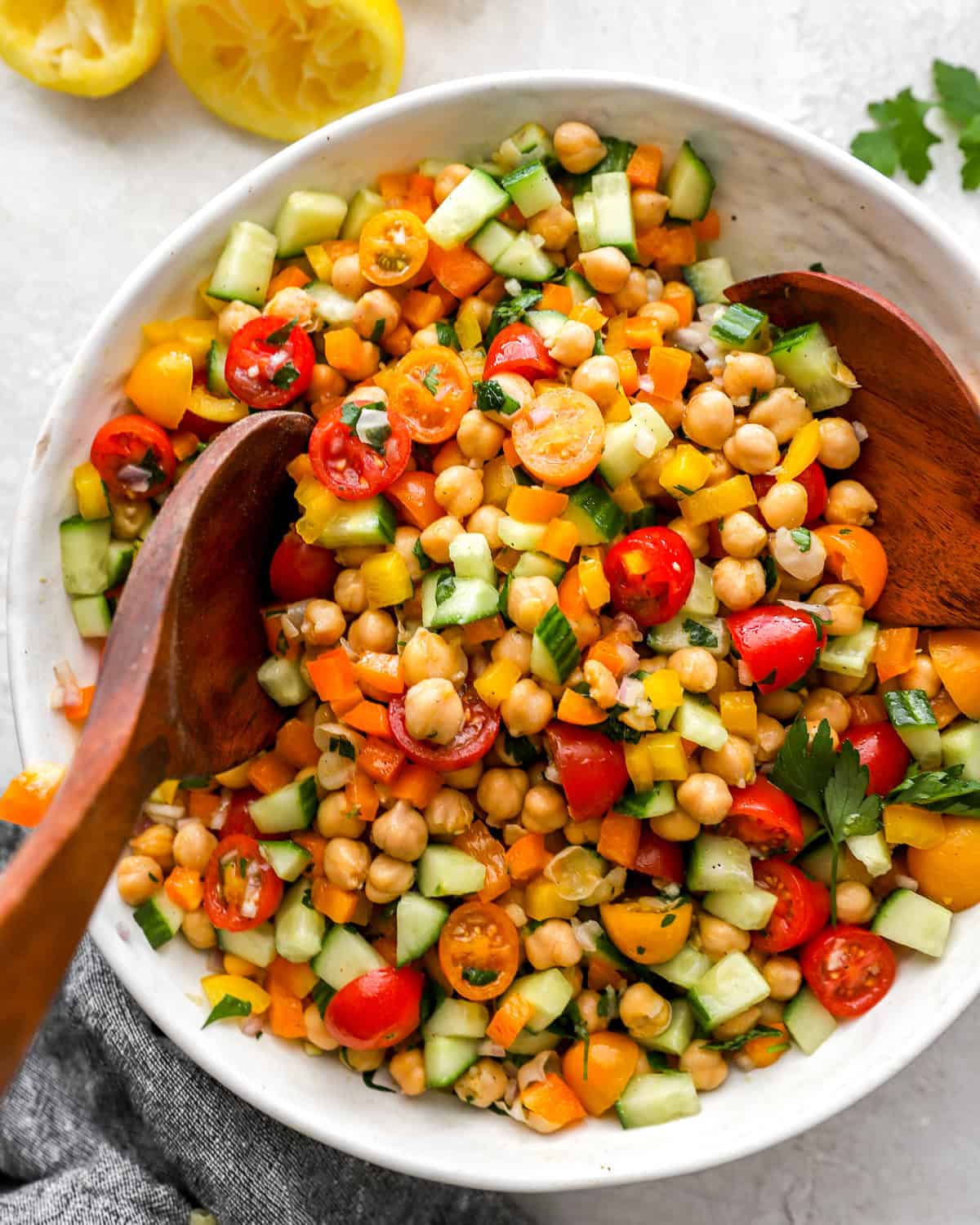 A large white bowl of colorful chickpea salad being mixed with wooden salad servers. The salad features chickpeas, chopped cucumber, halved cherry tomatoes, diced bell peppers, and fresh herbs.