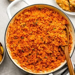 A large white pot filled with fluffy, vibrant orange jollof rice, garnished with chopped herbs, with a wooden spoon resting inside.