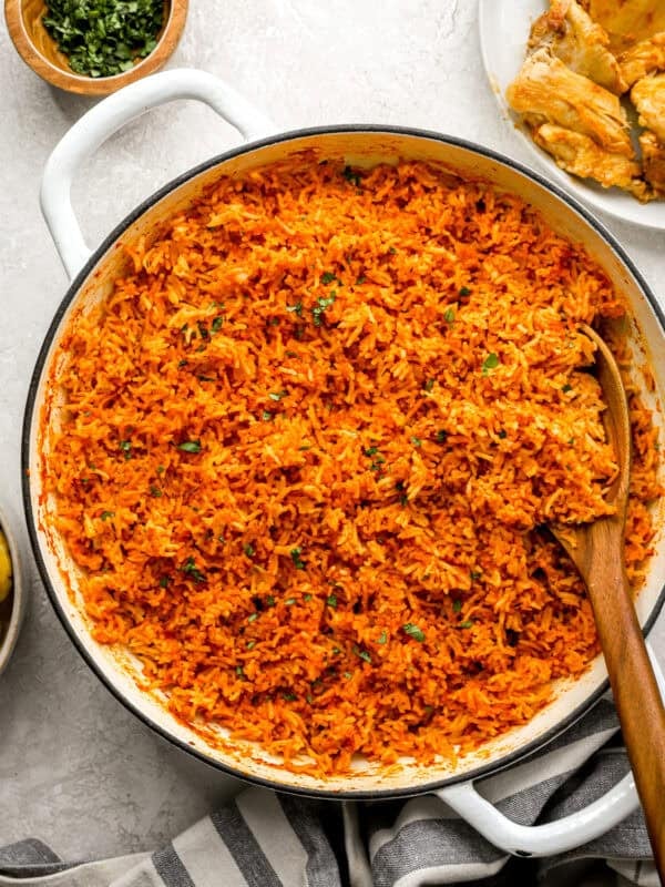 A large white pot filled with fluffy, vibrant orange jollof rice, garnished with chopped herbs, with a wooden spoon resting inside.