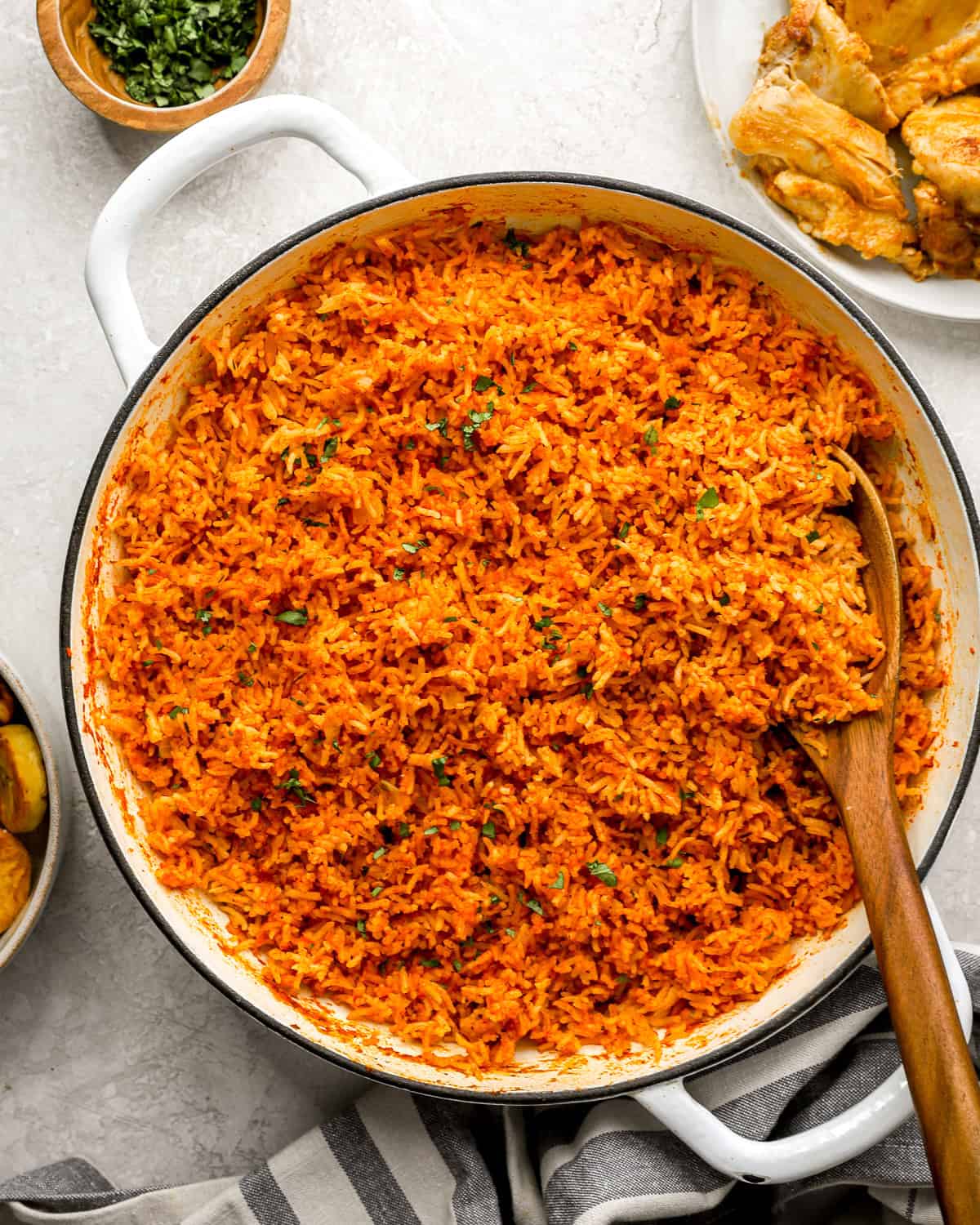 A large white pot filled with fluffy, vibrant orange jollof rice, garnished with chopped herbs, with a wooden spoon resting inside.
