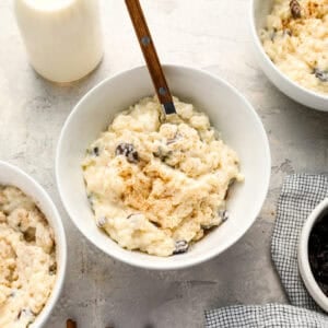A bowl of creamy rice pudding topped with cinnamon, with a wooden-handled spoon and a bottle of milk nearby, surrounded by other bowls of pudding and cinnamon sticks on a light countertop.