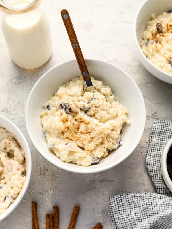 A bowl of creamy rice pudding topped with cinnamon, with a wooden-handled spoon and a bottle of milk nearby, surrounded by other bowls of pudding and cinnamon sticks on a light countertop.