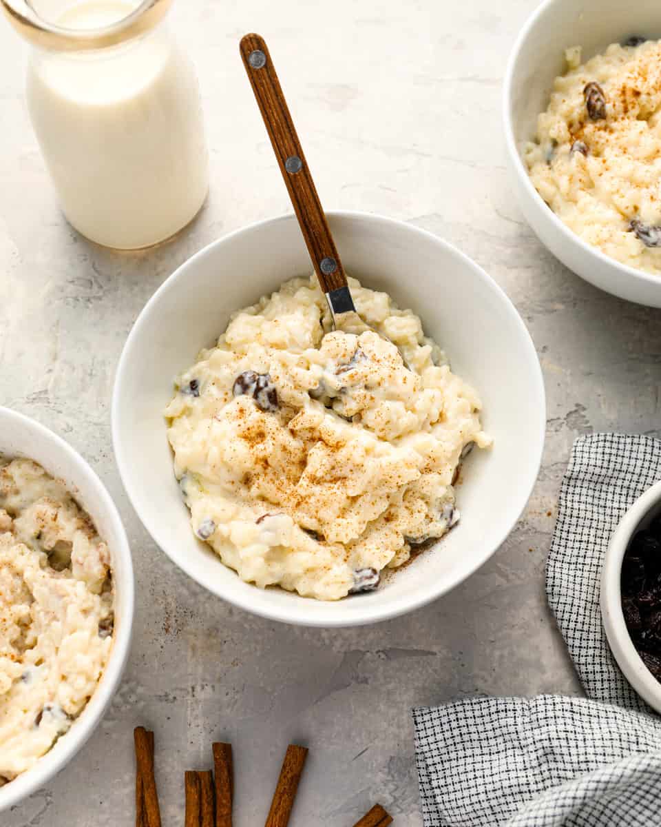 A bowl of creamy rice pudding topped with cinnamon, with a wooden-handled spoon and a bottle of milk nearby, surrounded by other bowls of pudding and cinnamon sticks on a light countertop.