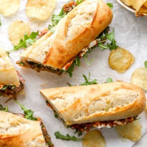 Toasted steak sandwiches filled with arugula, steak, and spreads, served on parchment with kettle chips scattered around.