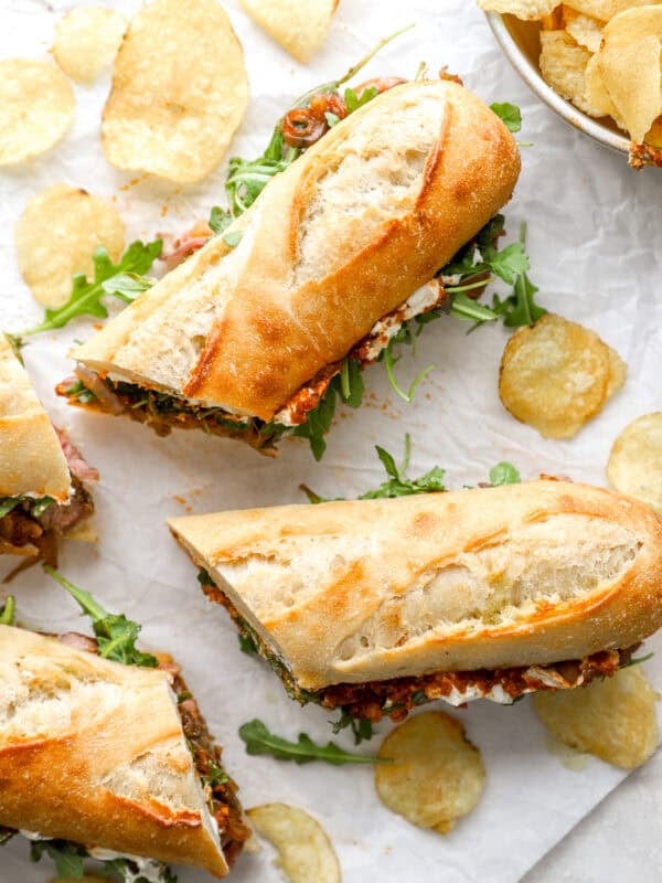 Toasted steak sandwiches filled with arugula, steak, and spreads, served on parchment with kettle chips scattered around.