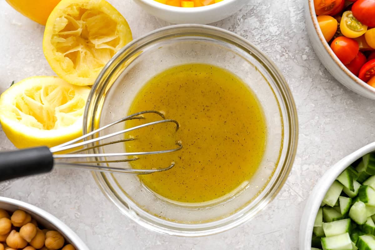 A glass bowl of golden lemon vinaigrette with a whisk resting inside. Freshly squeezed lemon halves, chickpeas, cucumbers, and cherry tomatoes surround the bowl.