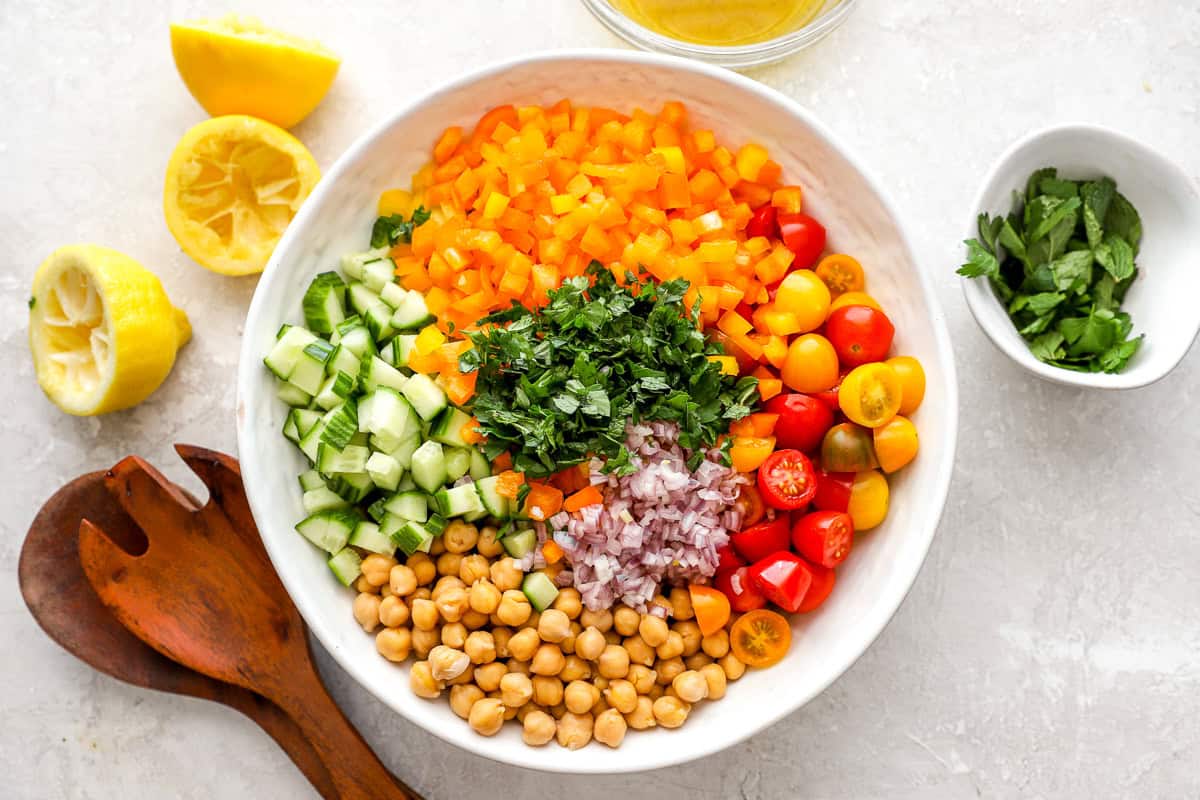 Ingredients for chickpea salad arranged in a large white bowl. Visible layers include chickpeas, chopped cucumber, diced yellow bell peppers, halved cherry tomatoes, finely chopped red onion, parsley, and mint.