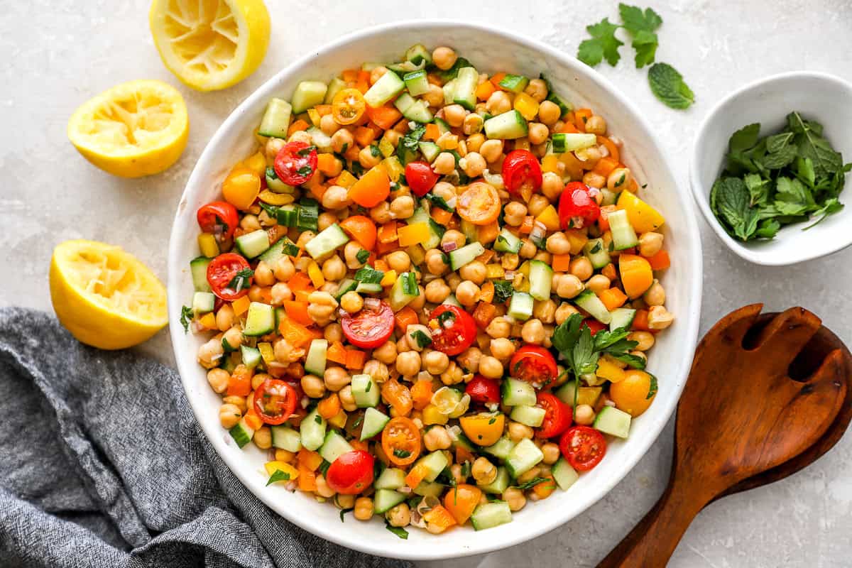 A top-down view of a finished chickpea salad in a white bowl surrounded by squeezed lemon halves, parsley, and a small dish of fresh mint.