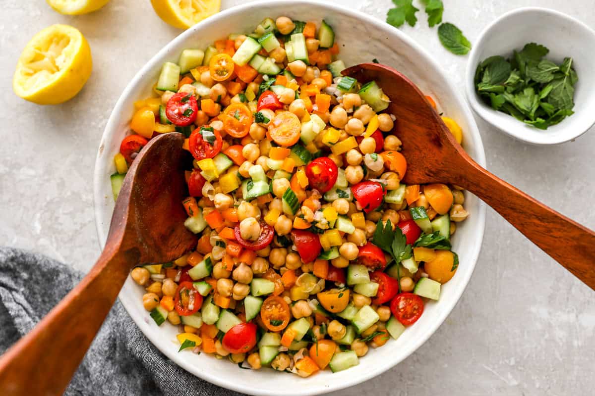 Chickpea salad in a white serving bowl with wooden utensils. The vibrant salad is surrounded by lemon halves and a small bowl of fresh mint.