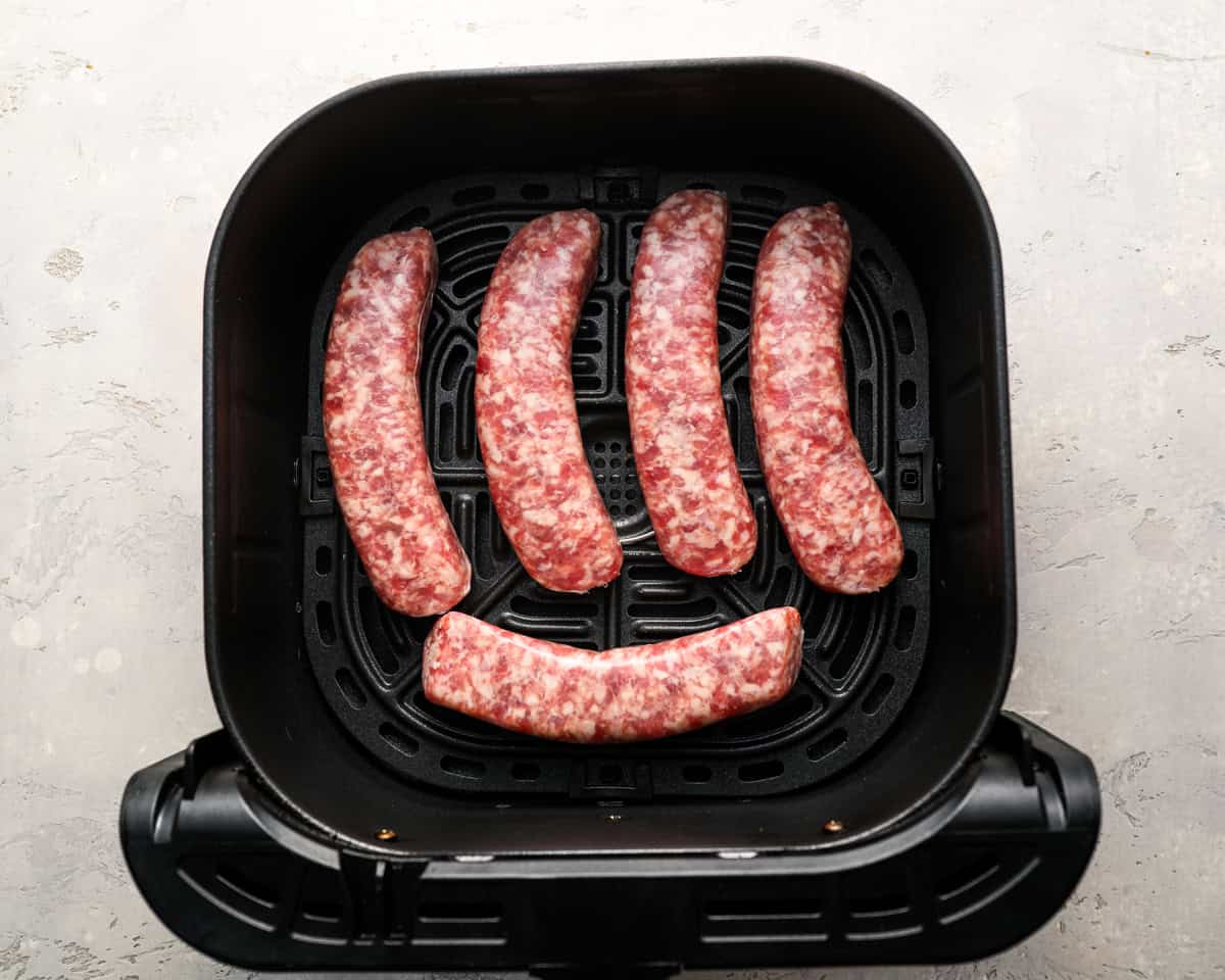 Uncooked bratwursts arranged in a single layer in an air fryer basket, spaced evenly apart.