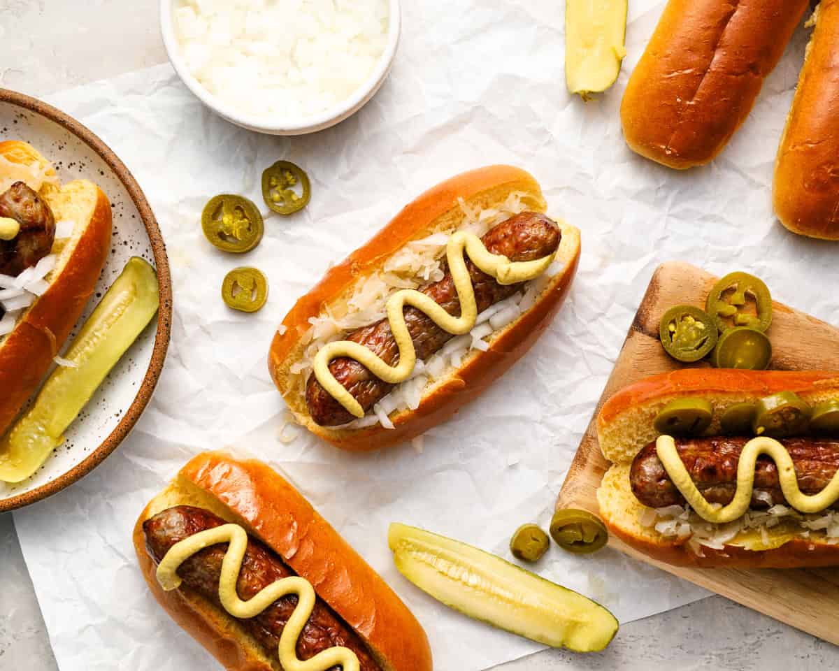 Overhead view of several fully assembled bratwurst buns with sauerkraut, mustard, jalapeños, and pickles, arranged on parchment paper with extra toppings scattered around.