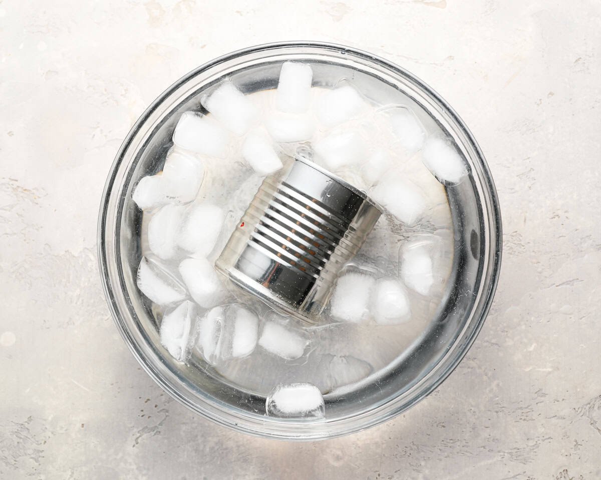 A can of caramel cooling in a bowl of ice water. The can is fully submerged with ice cubes floating around it.