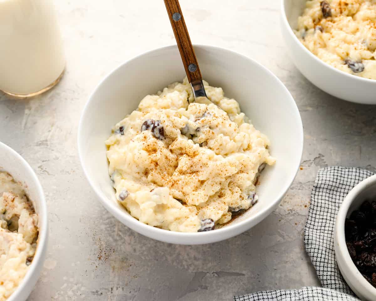 A close-up of a bowl of rice pudding with raisins and cinnamon, set next to a bowl of raisins, a bottle of milk, and a checked napkin, creating a cozy serving scene.