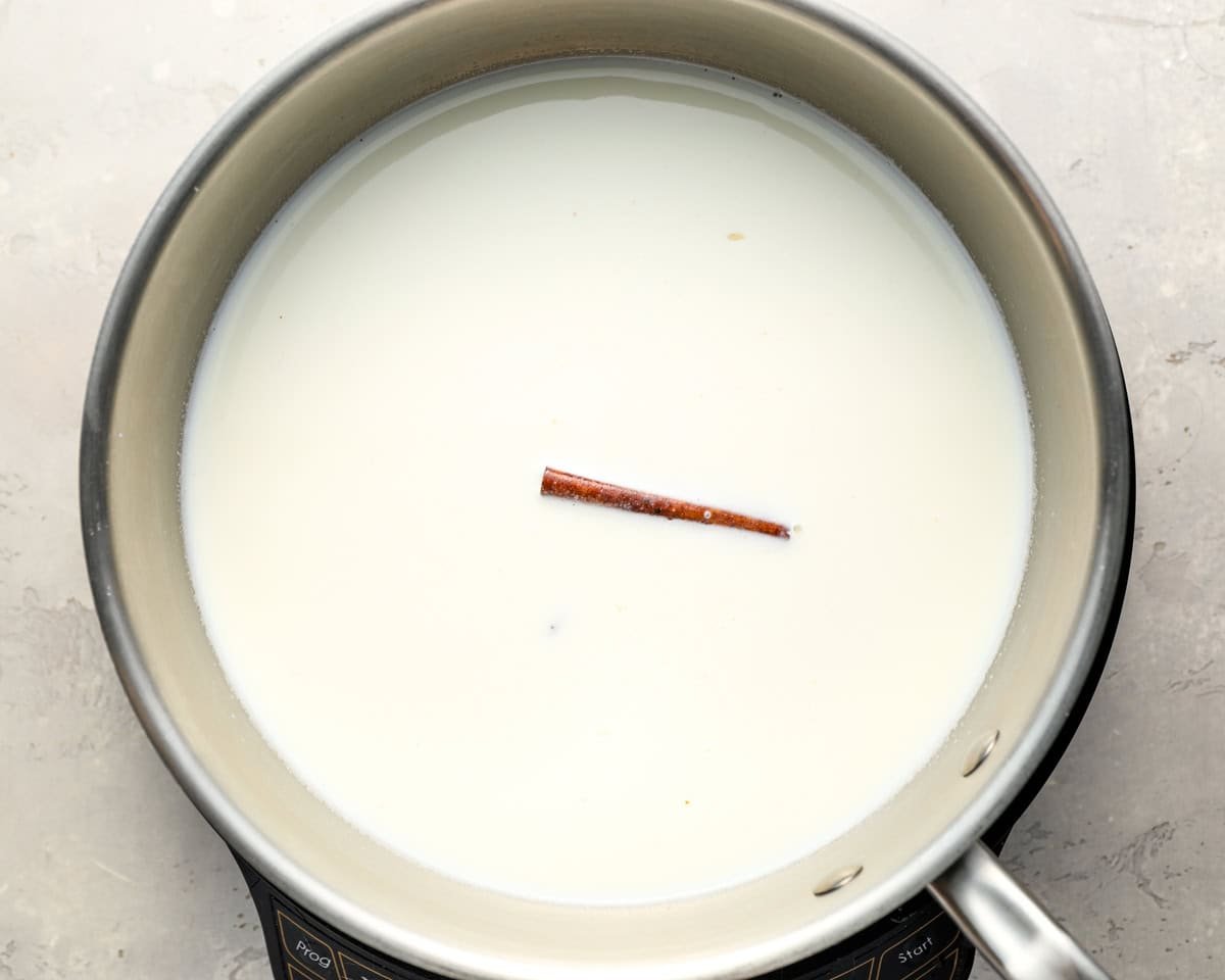 A saucepan filled with milk and a cinnamon stick floating in the center, set on a light-colored surface, showing the beginning stage of cooking rice pudding.