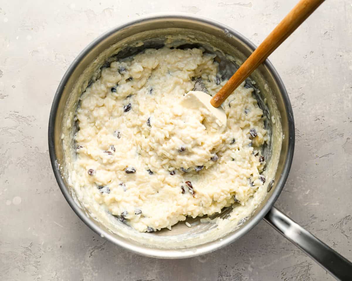 A pot of thick, creamy rice pudding being stirred with a white spatula, showing plump raisins throughout the mixture.