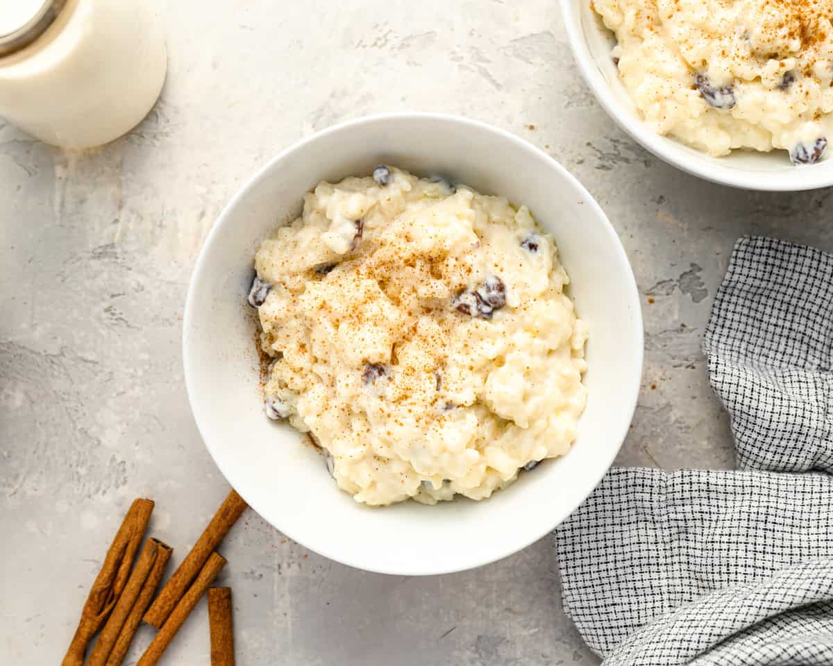 A bowl of rice pudding topped with cinnamon on a light background, with cinnamon sticks and a textured cloth napkin nearby.