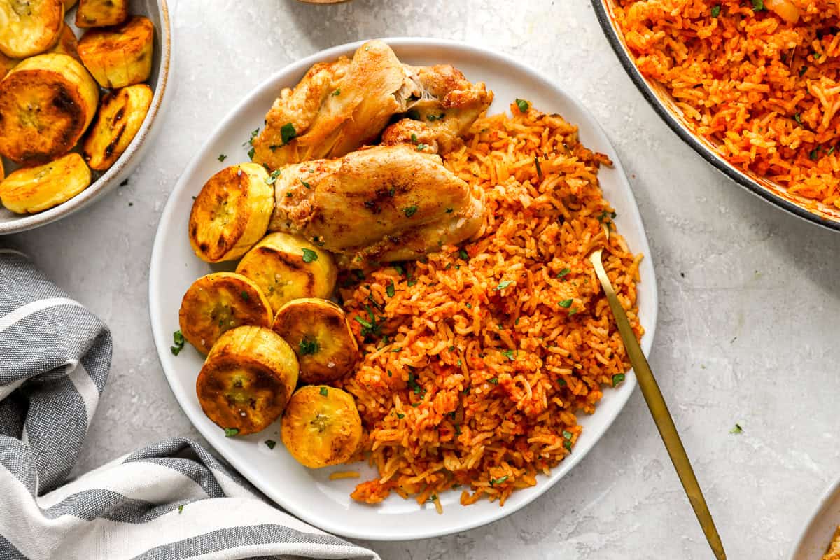 A plate of jollof rice served with golden fried plantains and seasoned chicken thighs, with a gold fork on the side.