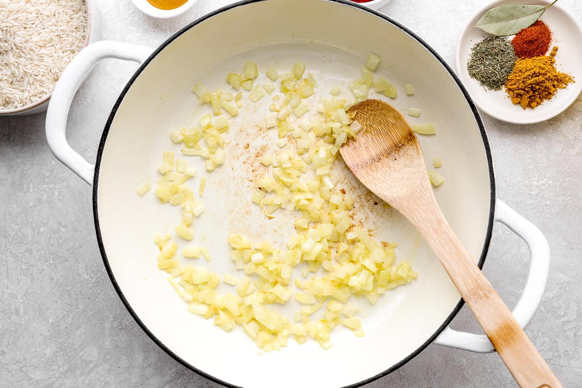 Diced onions sautéing in oil in a large white pot, with a wooden spoon and spices visible nearby.