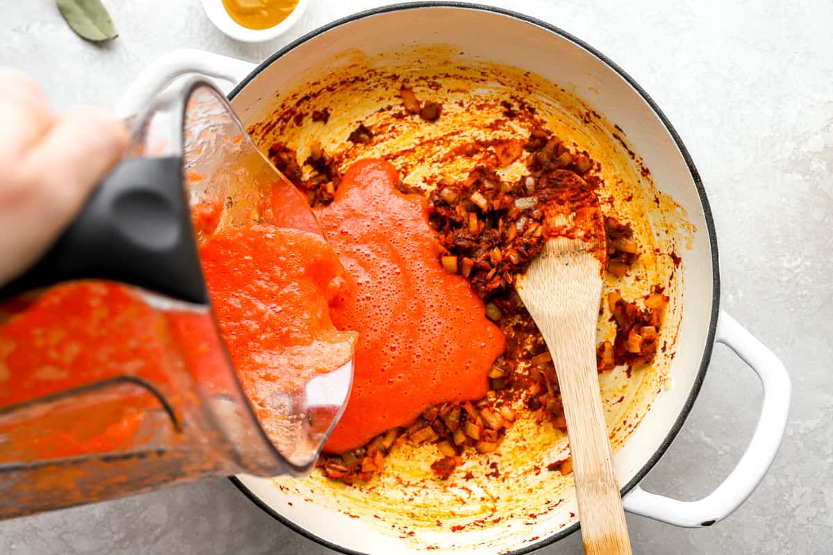 Tomato-pepper mixture being poured into a pot with sautéed onions and spices, starting the jollof rice base.