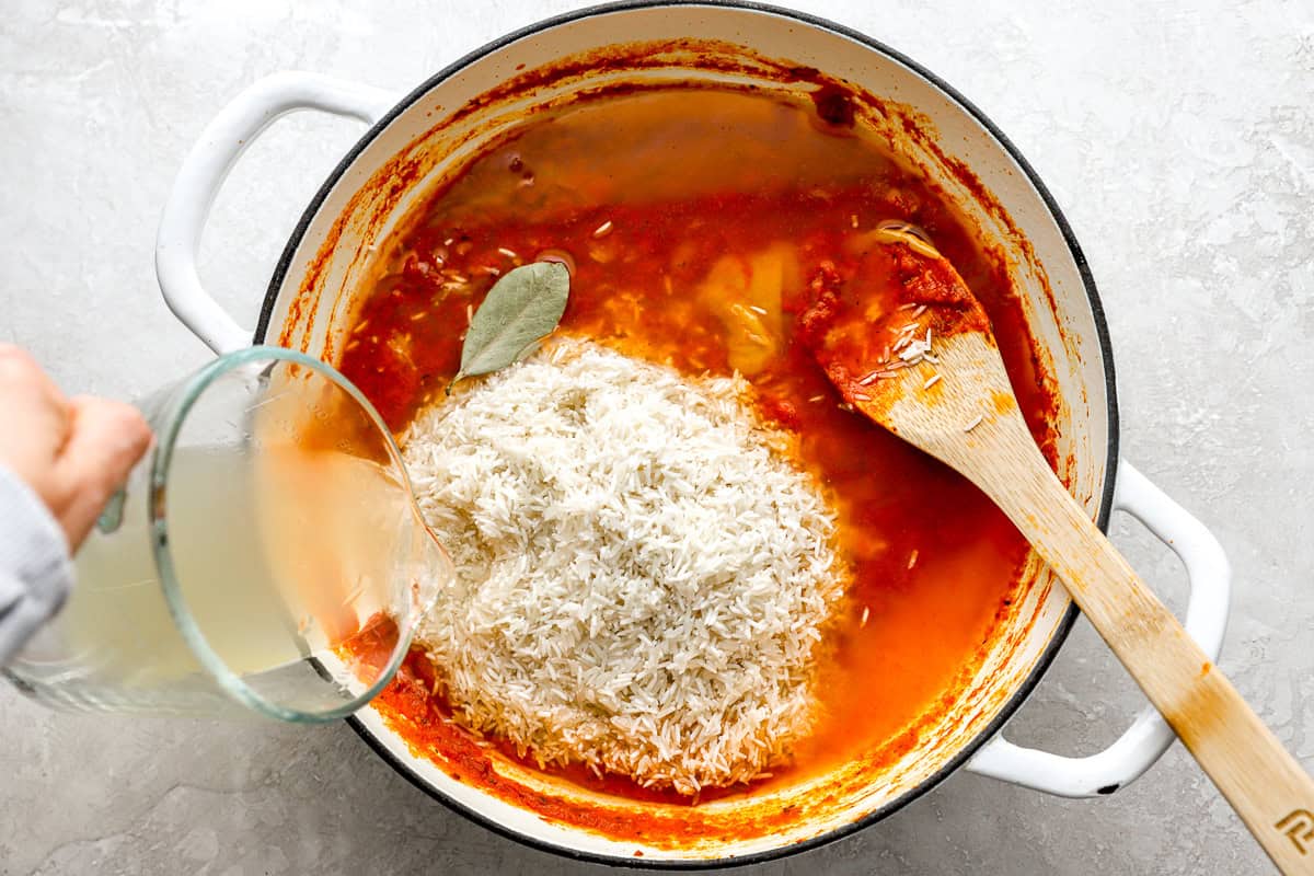 Uncooked rice and broth being added to the tomato mixture in a large pot, with a bay leaf floating on top.