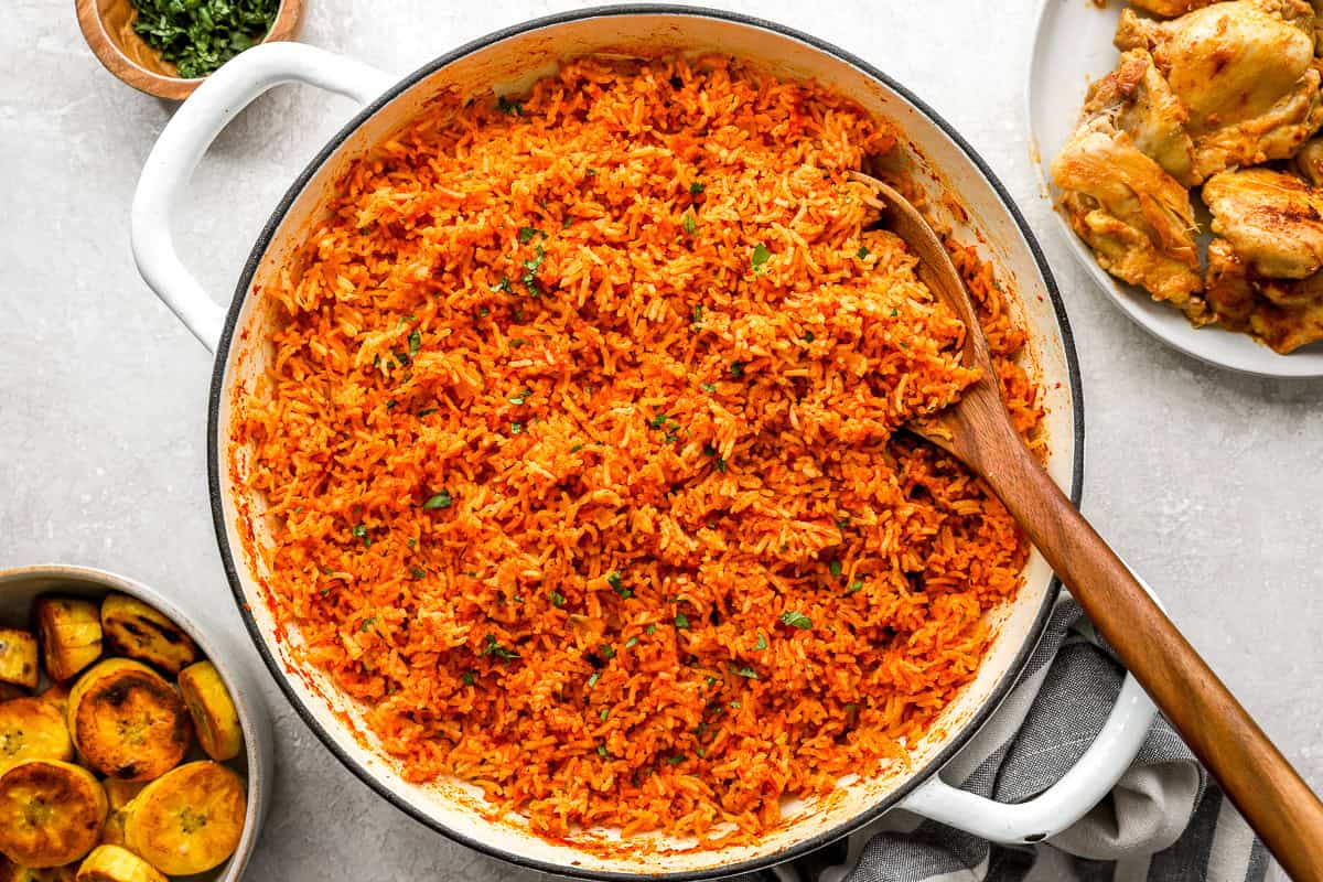 Overhead view of cooked jollof rice in a large white pot with a wooden spoon, surrounded by fried plantains and chicken.