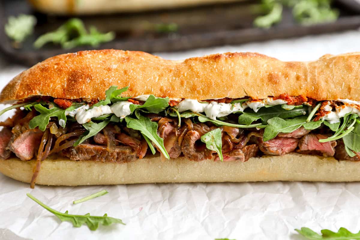Close-up of a fully assembled steak sandwich showing layers of arugula, garlic herb cream cheese, red pepper spread, caramelized onions, and sliced steak on crusty bread.