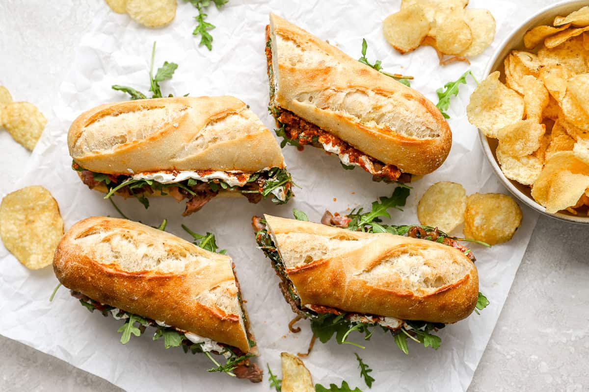 Four steak sandwiches cut in halves and arranged on parchment paper with kettle chips scattered around and in a bowl.