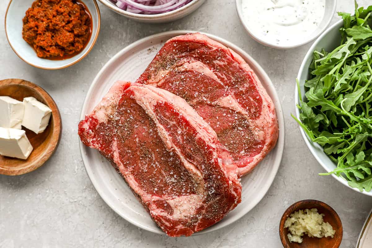 Seasoned raw ribeye steaks on a plate surrounded by bowls of garlic herb cream cheese, roasted red pepper spread, sliced red onions, minced garlic, and fresh arugula.