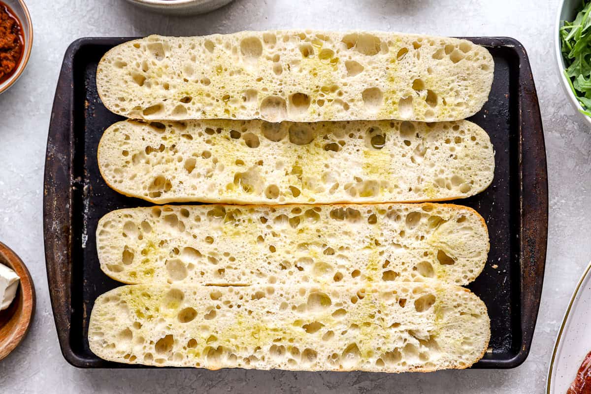 Sliced French baguettes drizzled with olive oil and placed on a baking sheet, ready to be toasted.