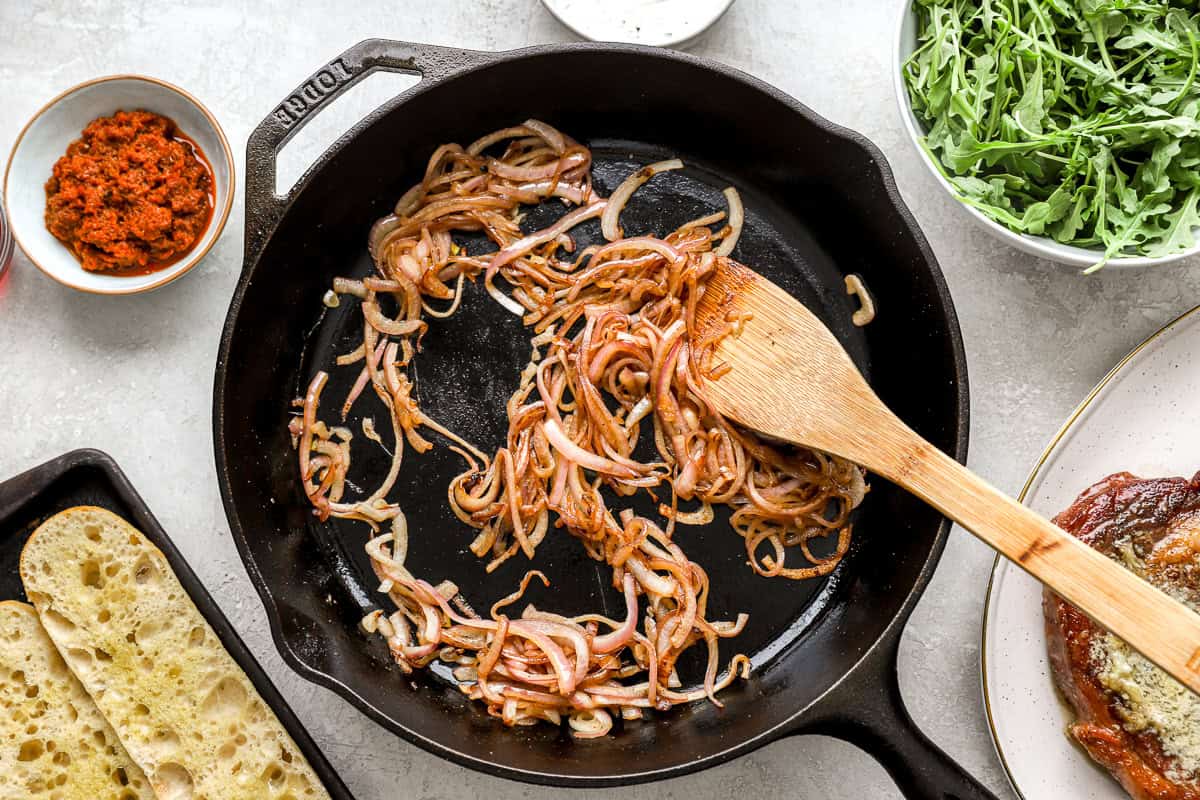 Sliced red onions being sautéed in a cast iron skillet with a wooden spatula.